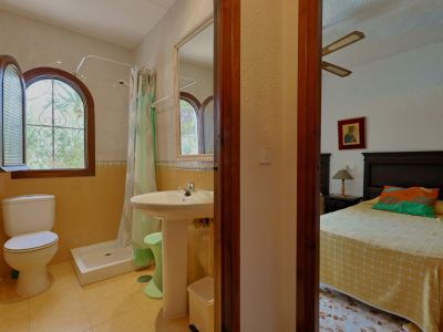 Salle de bain équipée d'une douche, toilettes et lavabo sur pied, avec une fenêtre arquée en bois donnant sur la végétation. Une ouverture mène à une chambre voisine.