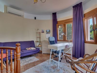 Bureau lumineux avec canapé violet, bureau en bois clair et chaise ergonomique. Grandes fenêtres en bois offrant une vue sur la verdure, climatisation et carrelage en terre cuite.