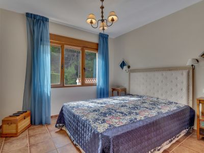 Chambre avec un grand lit à tête de lit capitonnée blanche, couvre-lit bleu à motifs et sol en terre cuite. Fenêtre en bois avec voilages bleus offrant lumière naturelle et vue sur la verdure.