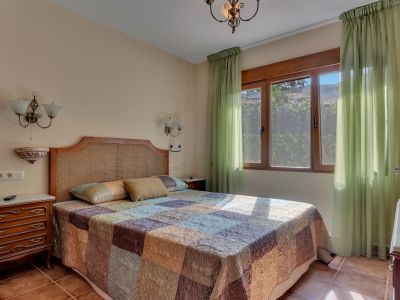 Chambre lumineuse avec tête de lit en rotin, courtepointe à motifs et grande fenêtre en bois avec rideaux verts transparents. Le sol en terre cuite et les tables de chevet classiques en bois.