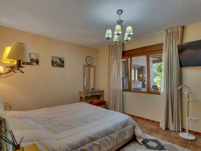 Chambre spacieuse avec grand lit, sol carrelé et fenêtre à cadre en bois donnant sur une zone couverte adjacente et le jardin. Comprend une coiffeuse et une télévision murale.