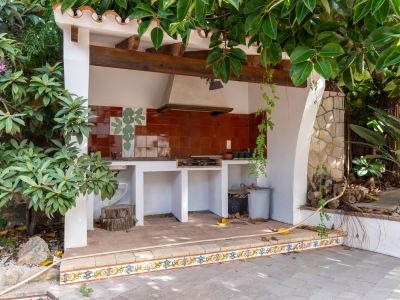 Cuisine extérieure avec barbecue intégré, crédence en carreaux de terre cuite à motif de cactus, et plafond à poutres en bois rustiques. La zone est carrelée et entourée de verdure luxuriante.