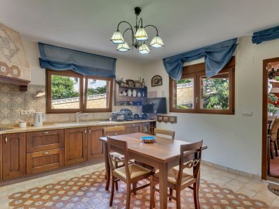 Cuisine fonctionnelle avec des armoires en bois foncé, un carrelage mural décoratif et un sol en carreaux à motifs. Deux fenêtres donnent sur le jardin, avec une table à manger en bois.
