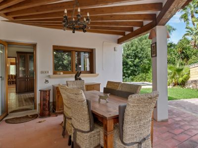 Espace repas extérieur couvert avec plafond à poutres en bois, sol en terre cuite et lustre en fer forgé. Des chaises en osier entourent une table en bois, donnant sur un jardin verdoyant.