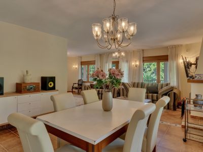 Espace salle à manger élégant avec lustre classique et chaises crème, ouvert sur un salon lumineux avec canapés rayés et grandes fenêtres donnant sur la verdure.
