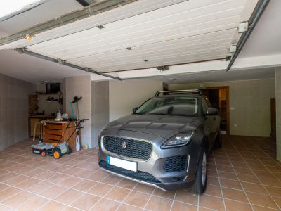 Garage privé avec sol carrelé en terre cuite et murs en carreaux gris. Cet espace fonctionnel comprend un établi avec outils, une tondeuse et une porte sectionnelle ouverte.