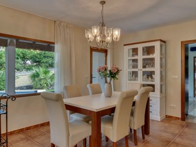 Salle à manger lumineuse avec table en bois, chaises rembourrées crème et lustre. Une grande fenêtre offre une vue sur le jardin luxuriant et la piscine privée.