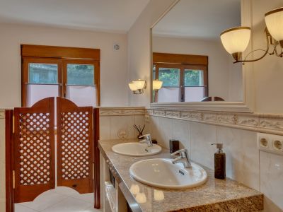 Salle de bain avec double vasque en granit, grand miroir encadré et appliques murales. Fenêtres en bois et portes battantes en treillis distinctives.