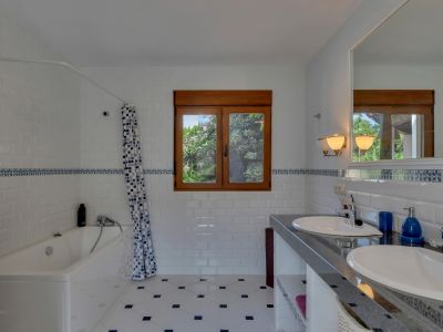 Salle de bain avec double vasque sur comptoir sombre et grand miroir. Carrelage métro blanc avec frise décorative bleue, sol à motifs et fenêtre en bois donnant sur la verdure.