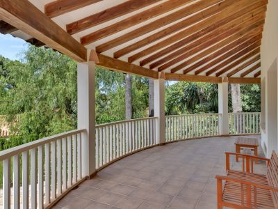 Terrasse couverte incurvée avec carrelage clair et plafond à poutres apparentes en bois. Une balustrade blanche encadre la vue sur la verdure luxuriante, agrémentée de bancs en bois.