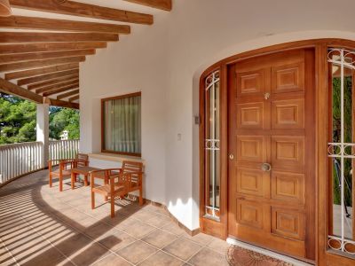 Terrasse d'entrée couverte avec porte en bois à panneaux, sol carrelé et poutres apparentes. Comprend un coin salon et une balustrade incurvée donnant sur la verdure.