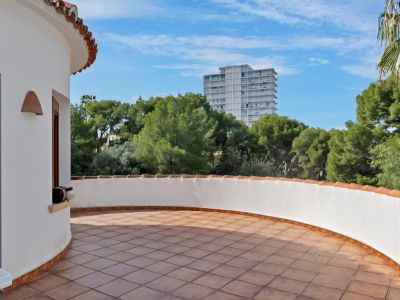 Terrasse extérieure incurvée avec carrelage en terre cuite et muret blanc surmonté de tuiles traditionnelles. Entourée d'arbres verts et d'un immeuble au loin sous un ciel bleu.