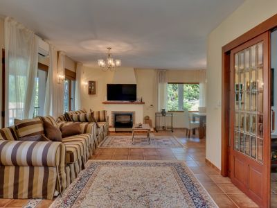 Vaste salon avec canapé rayé, cheminée et télévision. Le sol est en terre cuite, complété par de grandes fenêtres avec rideaux clairs et une porte coulissante en bois vitrée.
