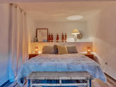 Chambre à coucher avec lit douillet, banc en rotin tressé et tables de chevet en bois. Une étagère au-dessus du lit présente des objets décoratifs et des lampes.