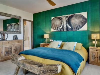 Chambre avec mur d'accent vert texturé, peinture d'éléphant et mobilier en bois naturel, incluant un banc sculpté et un miroir à motifs. Ventilateur de plafond visible.