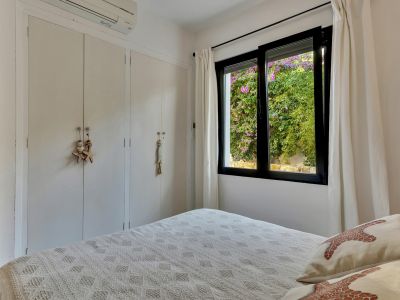 Chambre avec placards intégrés blancs, climatisation et fenêtre offrant une vue colorée sur la verdure luxuriante et les bougainvilliers roses.