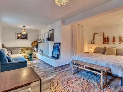 Chambre principale avec coin salon intégré, revêtement de sol imitation bois et tapis rond tressé. Comprend un lit avec banc en rotin, un canapé d'angle et une étagère murale.