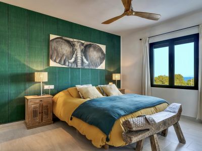Chambre spacieuse avec mur d'accent vert texturé, tableau d'éléphant et banc en bois sculpté. Fenêtre avec vue lointaine sur la mer et ventilateur de plafond en bois.