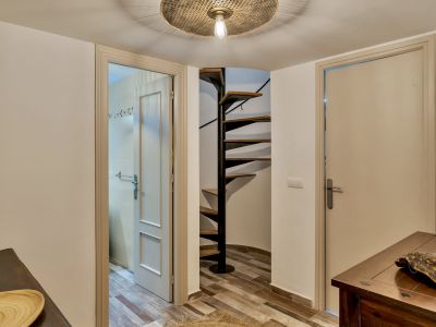 Couloir avec un escalier en colimaçon distinctif aux marches en bois et structure métallique noire, et un sol carrelé imitation bois. Deux portes sont visibles, dont une ouverte sur une salle de bain carrelée.
