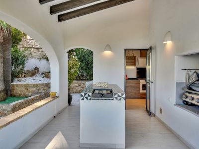 Cuisine extérieure couverte avec îlot central carrelé et plaque de cuisson intégrée, complétée par un barbecue encastré et des vues arquées sur le jardin palmier.