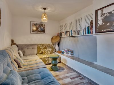 Espace salon confortable avec canapé en L, fenêtre hublot et bibliothèque intégrée sous des panneaux à persiennes. Une suspension tressée éclaire le parquet en bois.