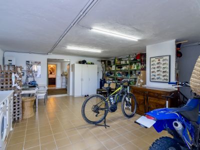 Grande buanderie et espace de rangement avec carrelage clair, comprenant une machine à laver, des étagères, un vélo électrique et une moto, éclairée par des néons au plafond.