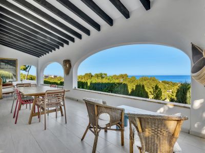 Grande terrasse couverte avec poutres en bois foncé et arches, offrant une vue panoramique sur la verdure luxuriante et la mer Méditerranée. Comprend un coin repas et des sièges en osier.