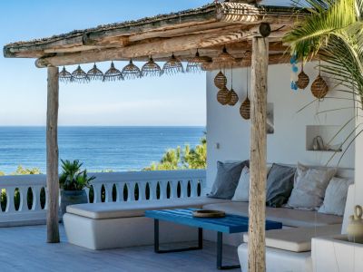 Pergola en bois rustique avec luminaires suspendus en osier, aménagée avec des banquettes blanches intégrées et une table basse bleue, dominant la mer.
