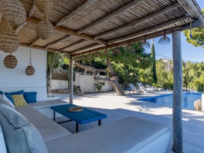 Pergola rustique avec toit de chaume et suspensions tressées, abritant un canapé extérieur avec coussins. La piscine bleue et la végétation luxuriante sont visibles en arrière-plan.