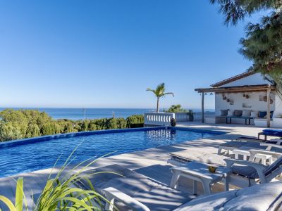 Piscine à débordement avec vue panoramique sur la mer, bordée de chaises longues et d'un coin salon rustique ombragé.