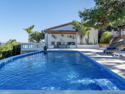 Piscine privée carrelée de mosaïque bleue avec vue sur la mer, adjacente à un salon extérieur ombragé par une pergola en bois.