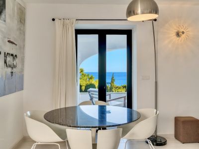 Salle à manger lumineuse avec table ronde en marbre foncé et chaises blanches, s'ouvrant sur une terrasse avec vue mer et verdure.