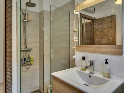 Salle de bain avec douche moderne à paroi vitrée, carrelage mural clair et sol en galets. Meuble-lavabo en bois clair et miroir encadré assorti.