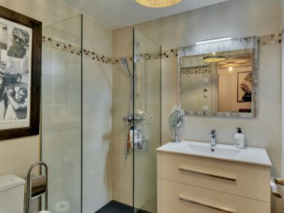 Salle de bain contemporaine avec douche à l'italienne vitrée, carrelage beige et frise mosaïque. Meuble-lavabo blanc et miroir encadré de bois patiné.