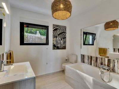 Salle de bain lumineuse avec baignoire, meuble-lavabo moderne et fenêtres offrant une vue sur la verdure extérieure. Une suspension en osier et une œuvre d'art zébrée ajoutent une touche distinctive.