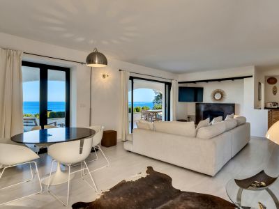 Salon et salle à manger ouverts avec une table ovale foncée, des chaises blanches et un canapé clair, s'ouvrant sur une terrasse avec vue panoramique sur la mer.