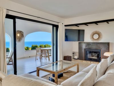 Salon lumineux avec canapé clair, table basse en bois et cheminée moderne. De grandes baies vitrées s'ouvrent sur une terrasse offrant une vue mer panoramique.