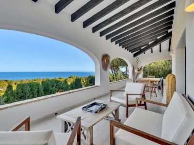 Terrasse couverte avec poutres en bois foncé et carrelage blanc. Elle offre une vue panoramique sur la mer à travers des arches, meublée de sièges extérieurs en bois.