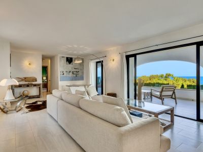 Vaste salon avec canapé beige, carrelage clair et accès direct à une terrasse offrant une vue panoramique sur la mer et une végétation luxuriante. Table basse moderne et art mural.