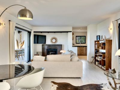 Vaste salon lumineux avec canapé clair, cheminée contemporaine et télévision intégrée. Des portes coulissantes mènent à la terrasse, complété par une table à manger et une bibliothèque en bois.