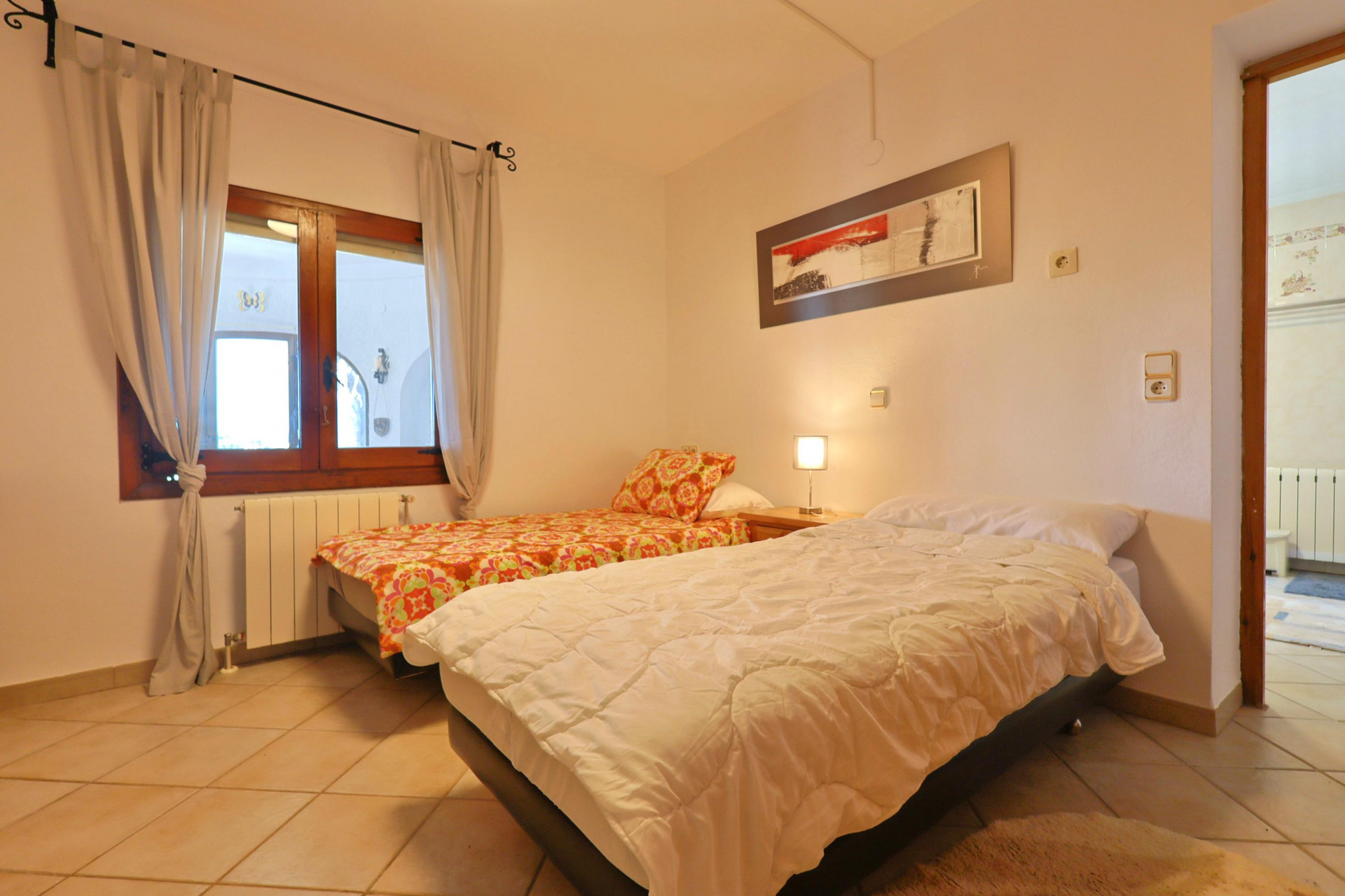 Chambre lumineuse avec lits jumeaux, fenêtre en bois et sol carrelé.