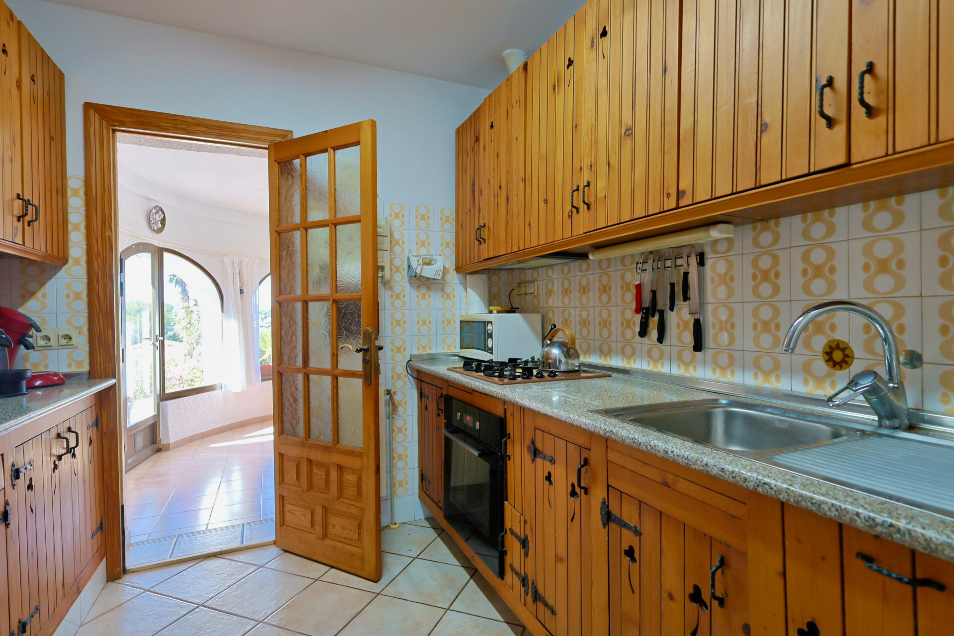 Cuisine rustique avec meubles en bois et accès à un salon lumineux avec de grandes fenêtres.