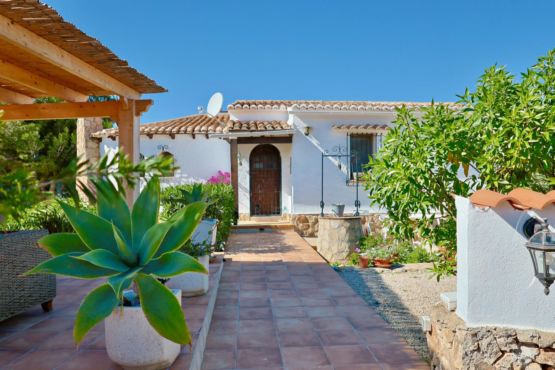 Entrée de la villa avec allée carrelée, verdure méditerranéenne et puits en pierre.