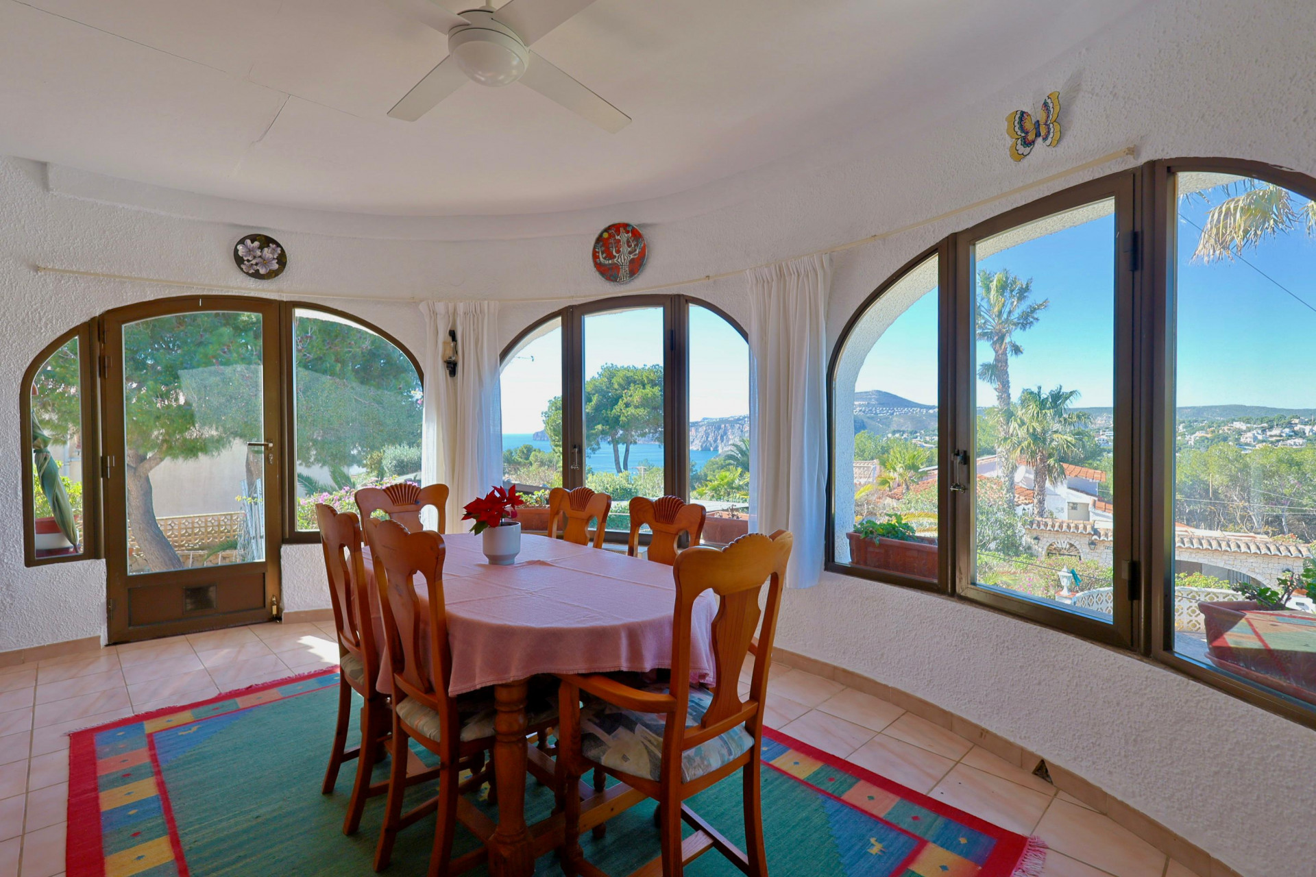 Salle à manger lumineuse avec vue sur mer par des fenêtres cintrées. Table en bois avec nappe rose.