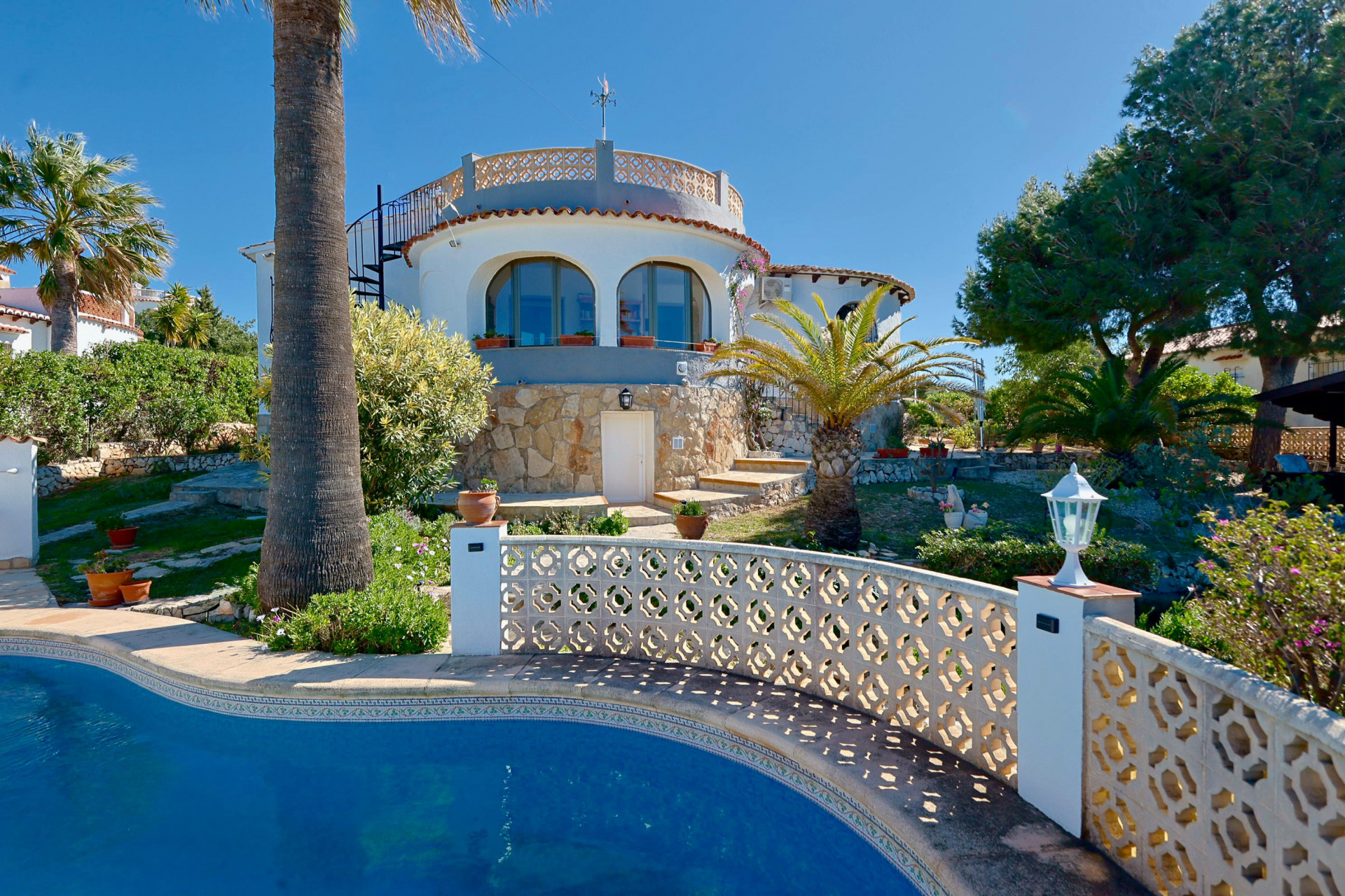Vue de la piscine avec rambarde à croisillons et façade en pierre de la villa à Jávea.