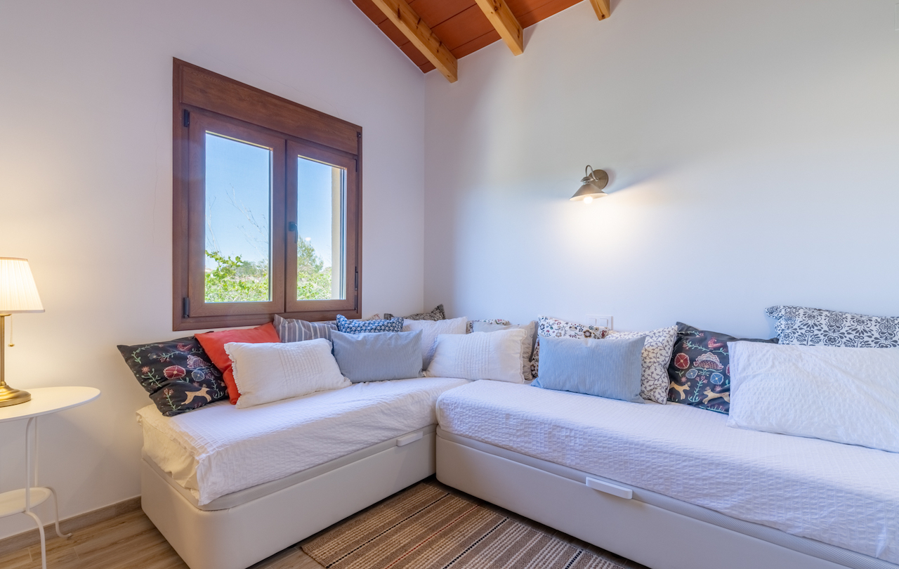 Chambre avec canapé-lit en L, plafond à poutres apparentes et fenêtre en bois donnant sur le jardin. L'espace est complété par un éclairage tamisé.