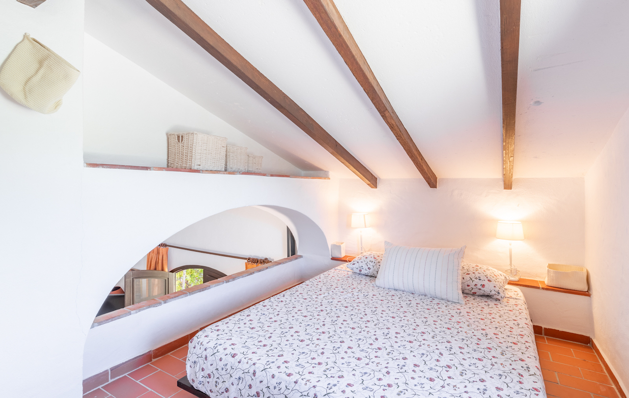 Chambre mansardée avec poutres en bois apparentes, sol en terre cuite et une ouverture en arche offrant une vue sur l'étage inférieur de la finca.