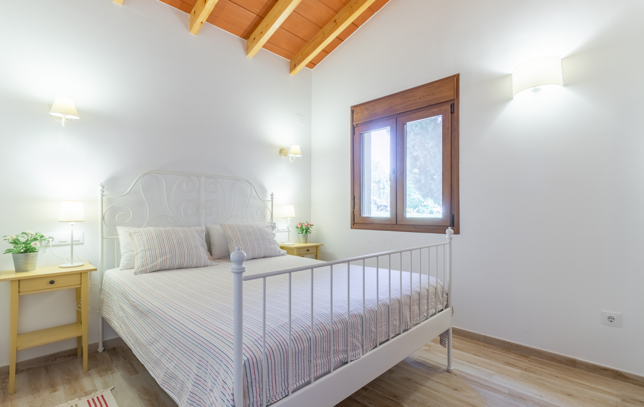 Chambre rustique avec plafond à poutres apparentes, lit en fer forgé blanc et fenêtre en bois apportant une lumière naturelle.