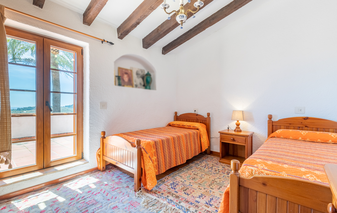 Chambre rustique avec poutres apparentes, lits jumeaux et niche murale décorative. Une porte-fenêtre en bois donne accès à une terrasse ensoleillée avec vue sur le jardin.
