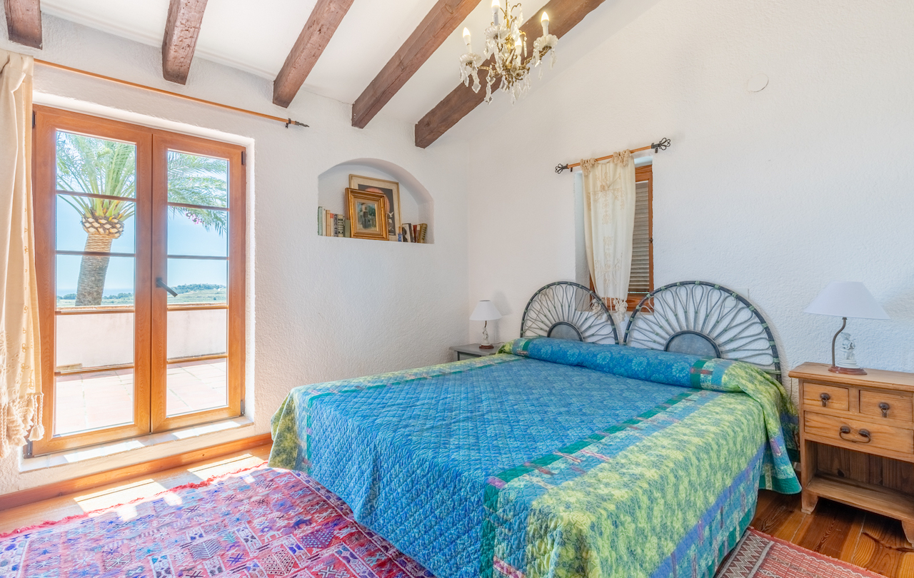 Chambre rustique avec poutres apparentes, niche murale décorative et accès direct à la terrasse offrant une vue sur les palmiers. Sols en bois et tête de lit en fer forgé.